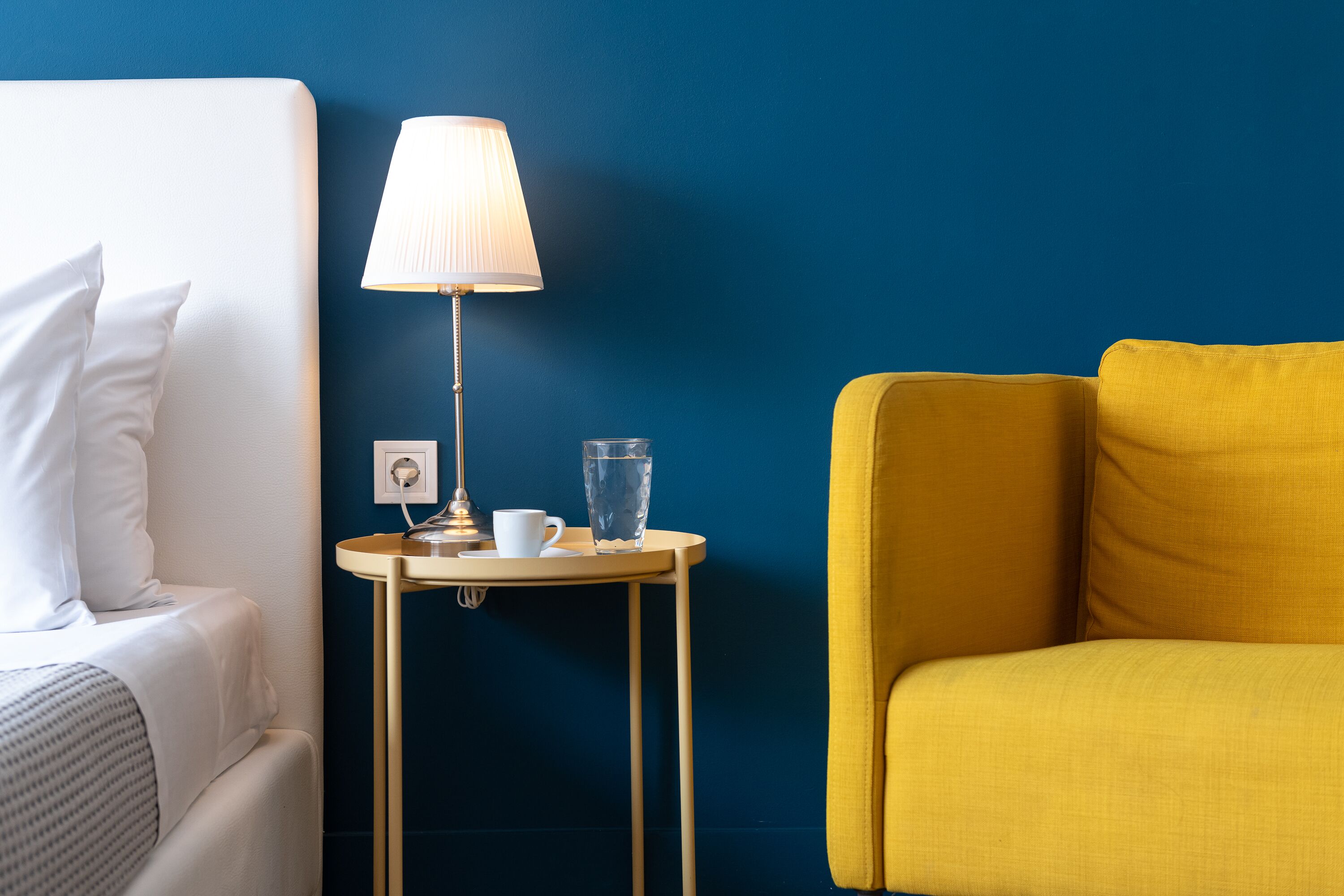 A hotel room with a bed, a bedside table with a lamp, a cup, and a glass of water.