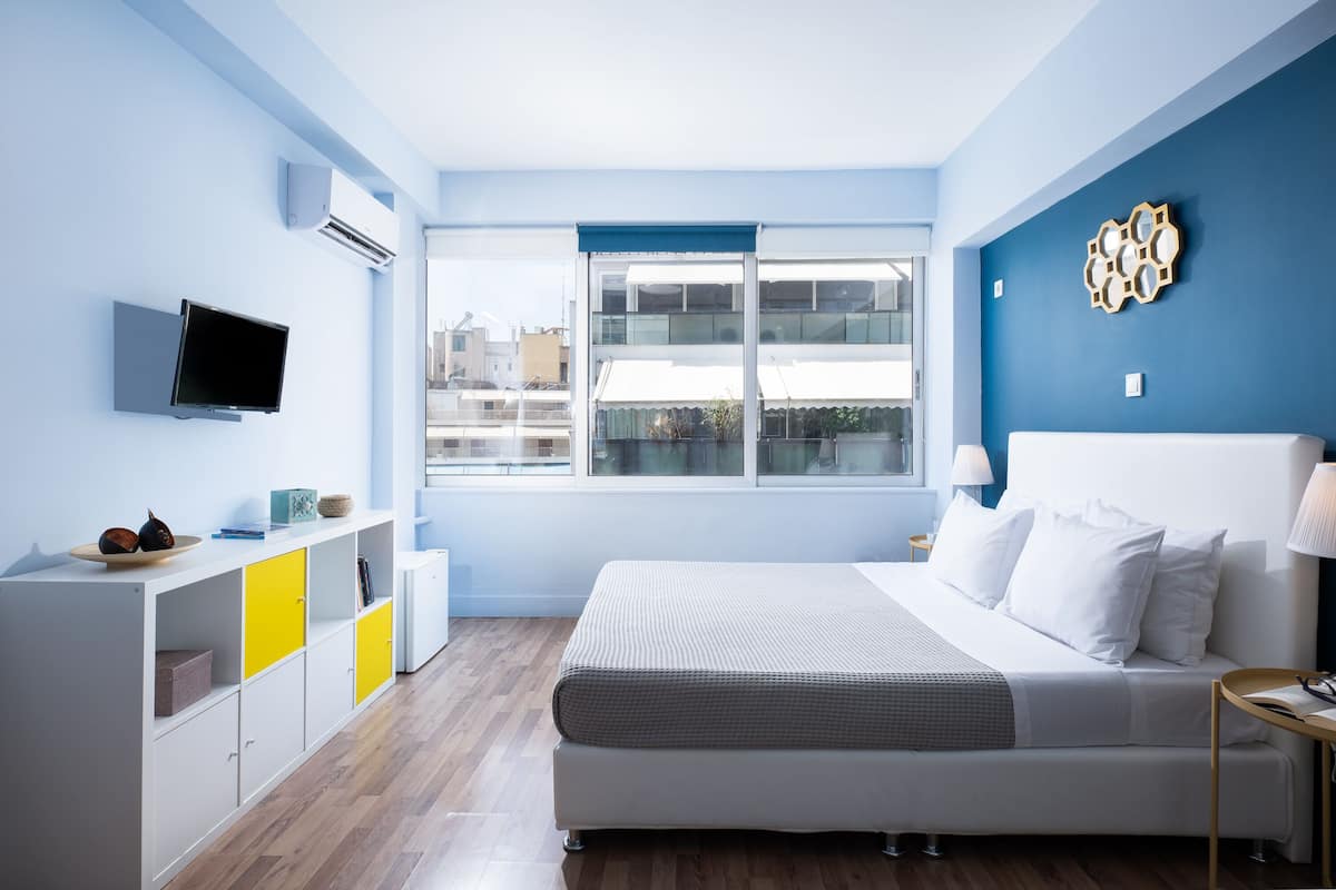 A modern bedroom with a large bed, a TV mounted on the wall, and a window with a view of buildings.