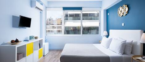 A modern bedroom with a large bed, a TV mounted on the wall, and a window with a view of buildings.