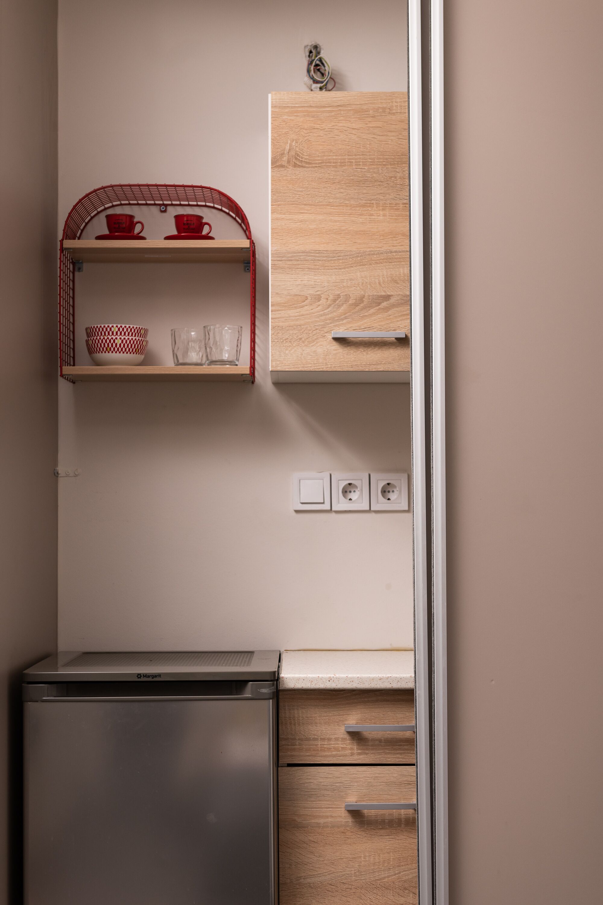 A compact kitchen with a stainless steel refrigerator, wooden cabinets, and a red wire shelf holding cups and glasses.