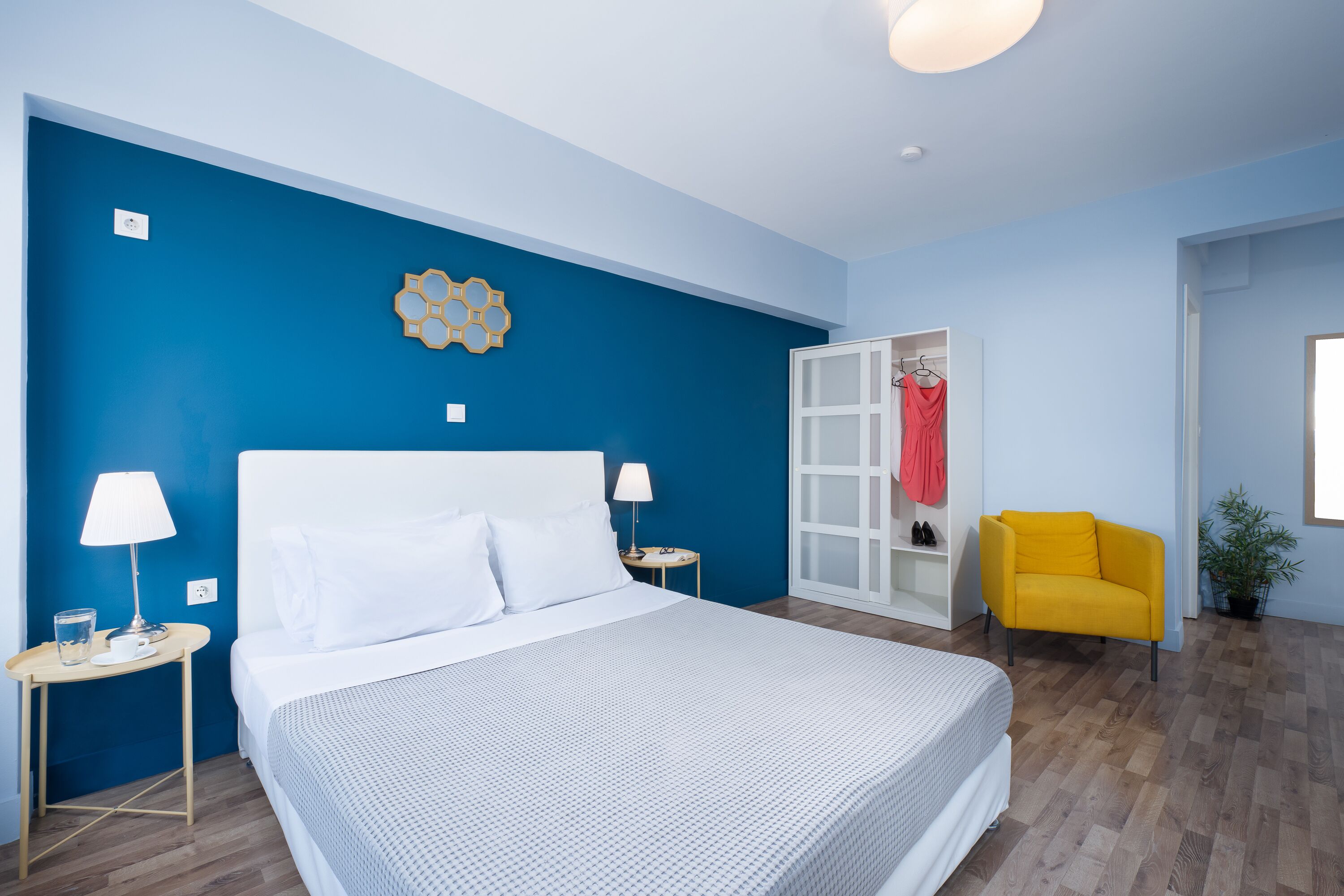 Una camera da letto con un letto grande, due comodini con lampade, una sedia giallo brillante, un armadio e una parete decorata in blu.