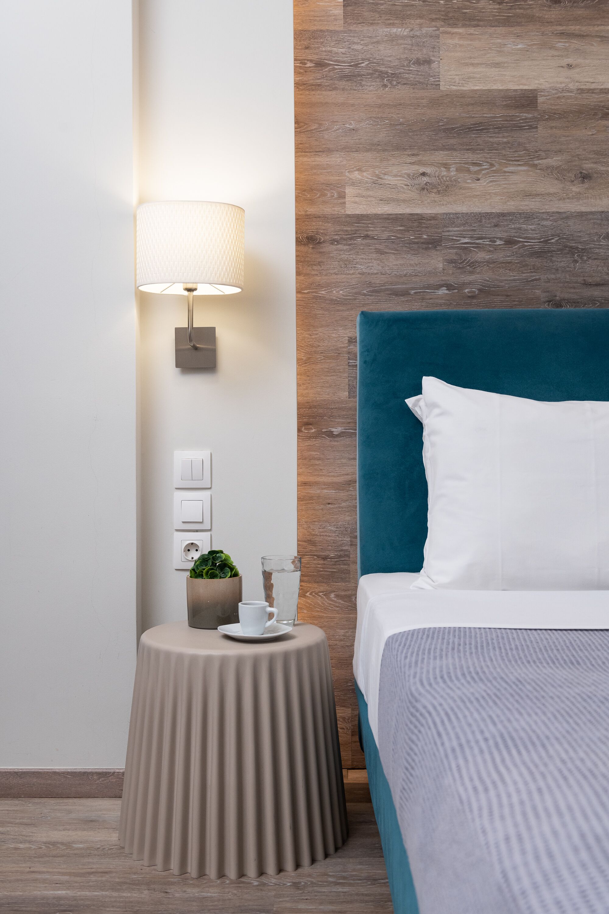 A hotel room with a bed, a bedside table with a lamp, a small potted plant, and a glass of water.