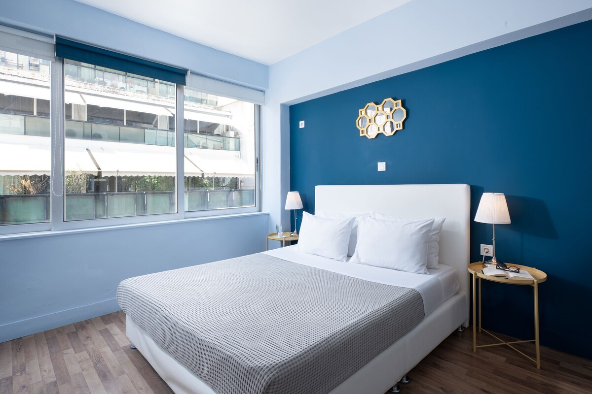 A modern hotel room with a large bed, two bedside tables, a window with a view of a building, and a blue accent wall.