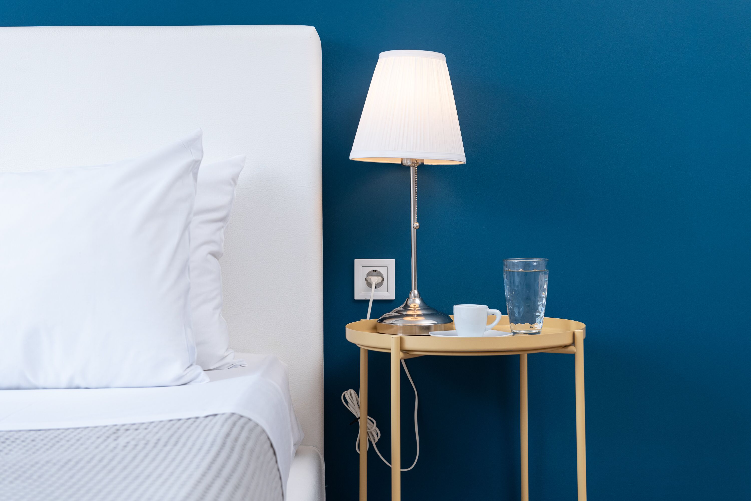 A neatly made bed with white bedding, a bedside table with a lamp, a cup, and a glass of water.