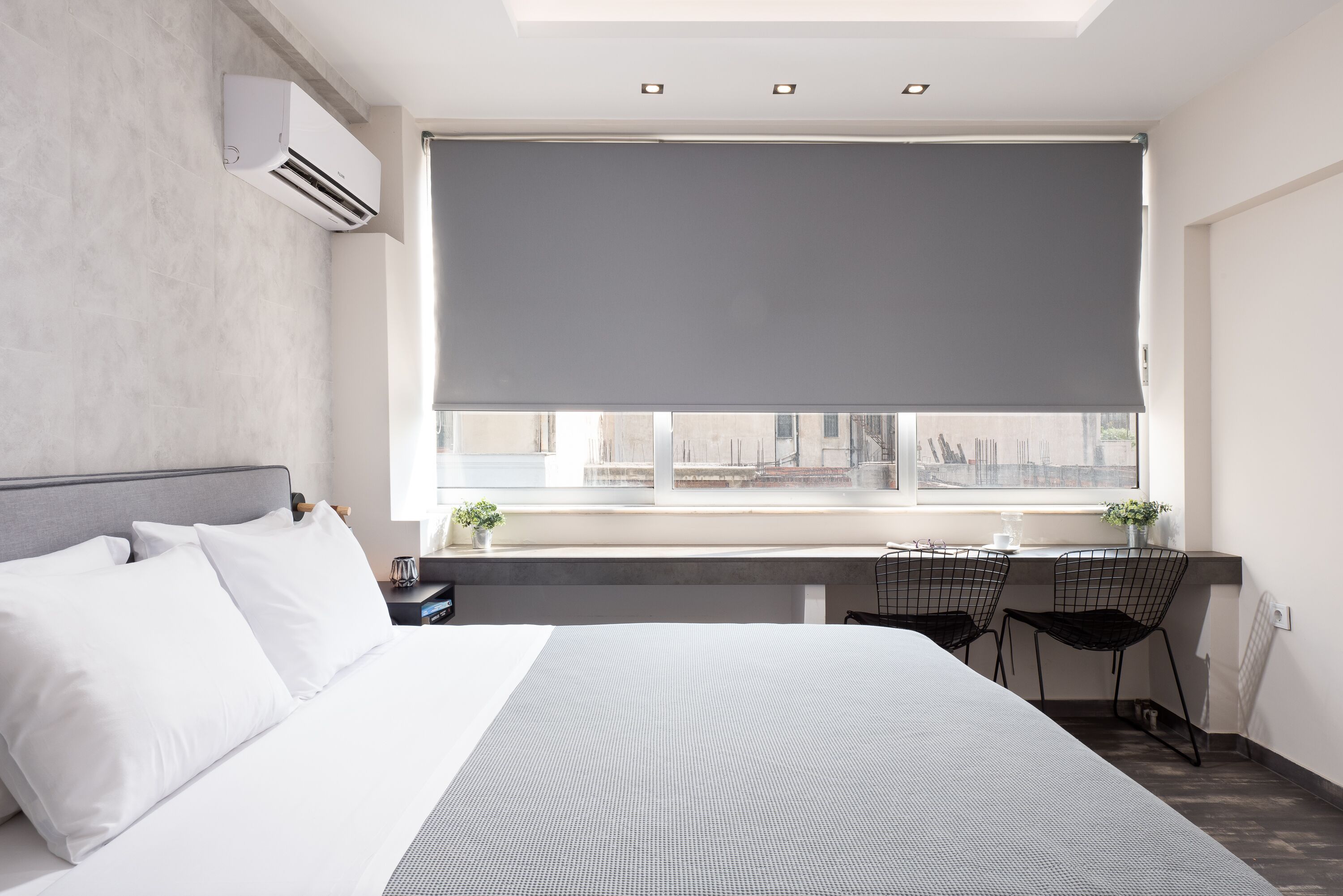 Una habitación de hotel moderna con una cama grande, un escritorio con dos sillas, un ventanal con persiana y un equipo de aire acondicionado en la pared.