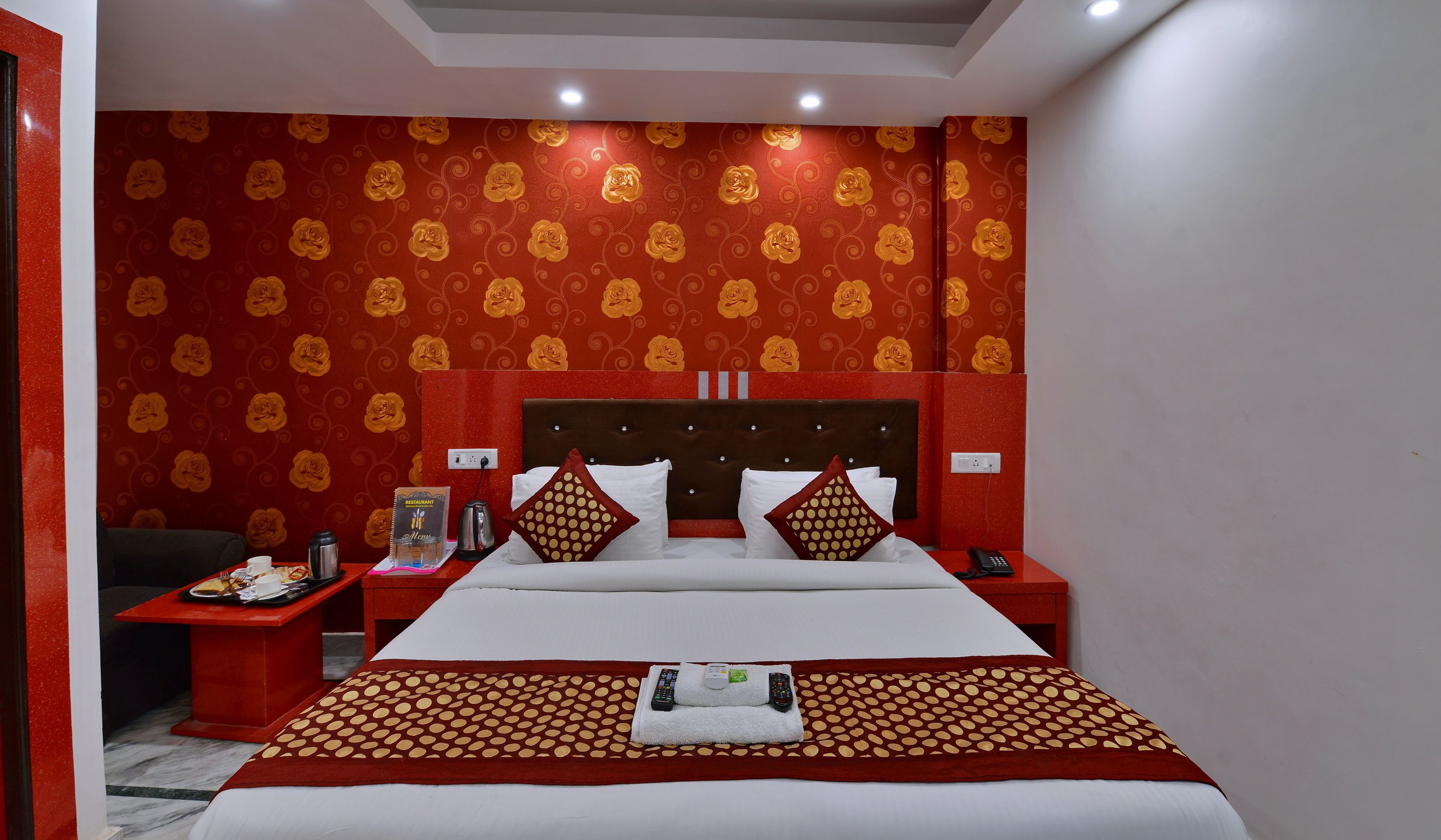 A hotel room with a large bed, red and gold patterned wallpaper, a red nightstand with a phone and a remote control, and a small table with a menu and a kettle.