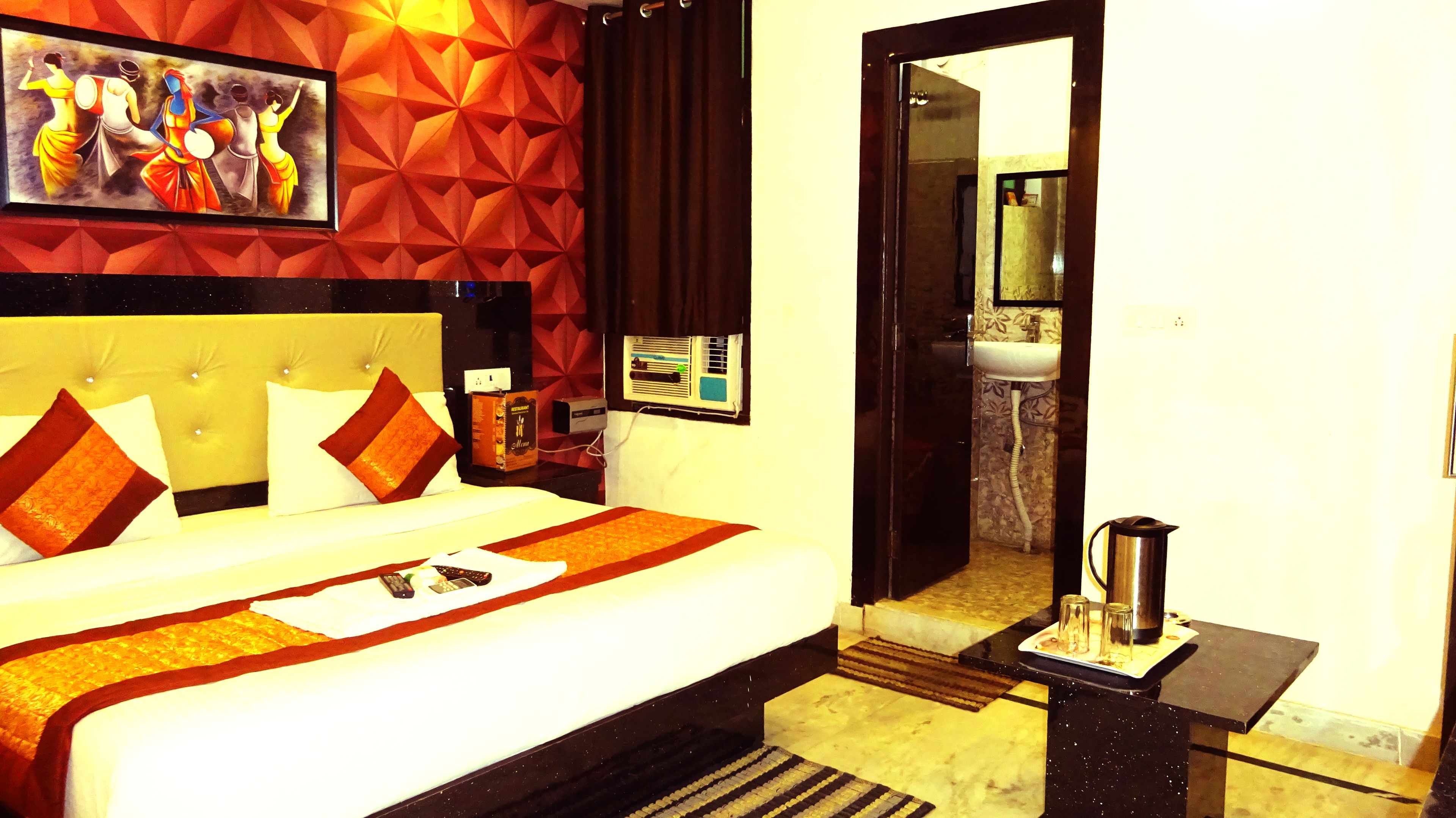 A hotel room with a large bed, a painting on the wall, a small table with a kettle, and a bathroom visible through an open door.