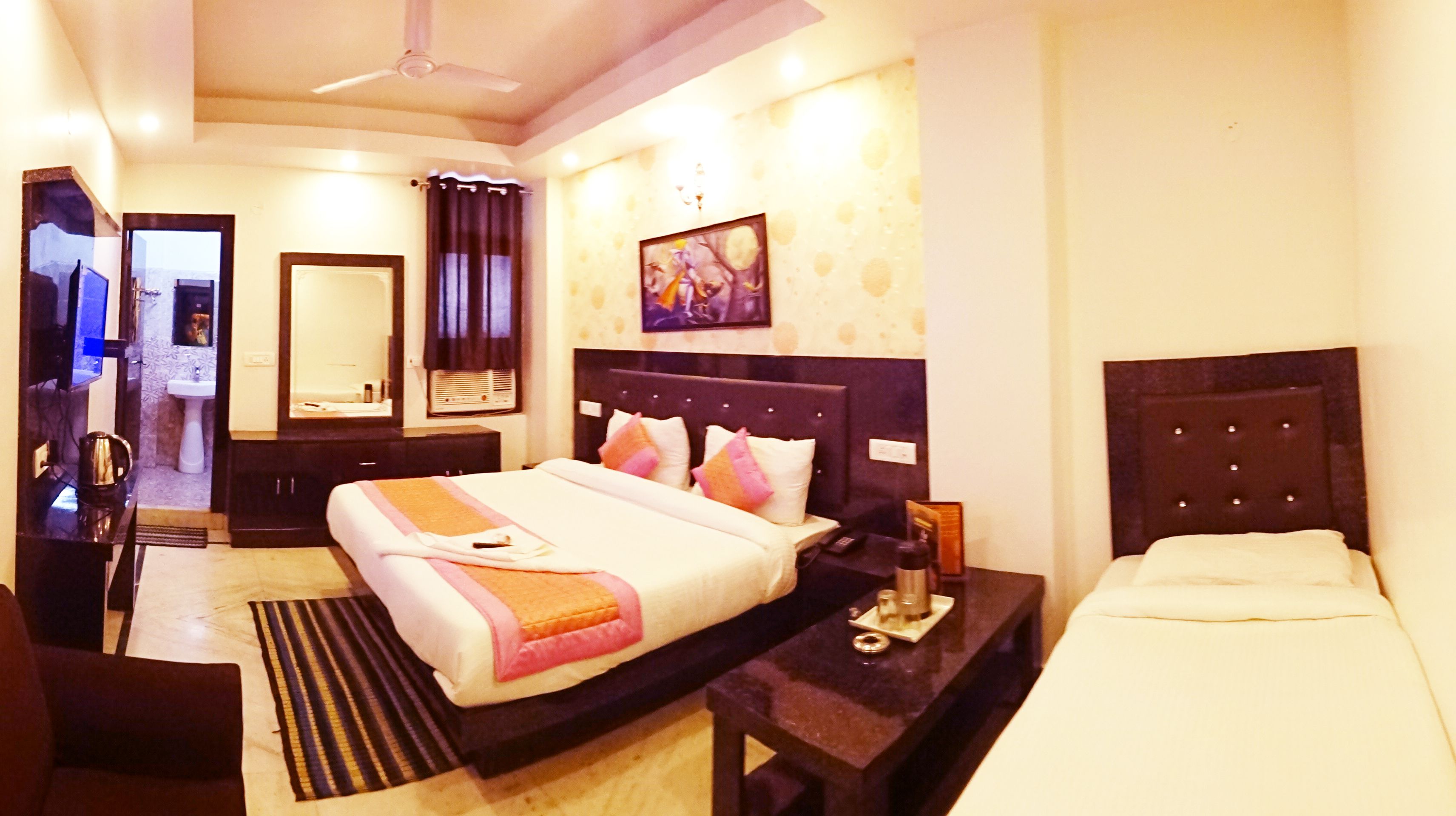 A hotel room with a large bed, bedside tables, a desk, a chair, a mirror, a painting on the wall, and a ceiling fan.