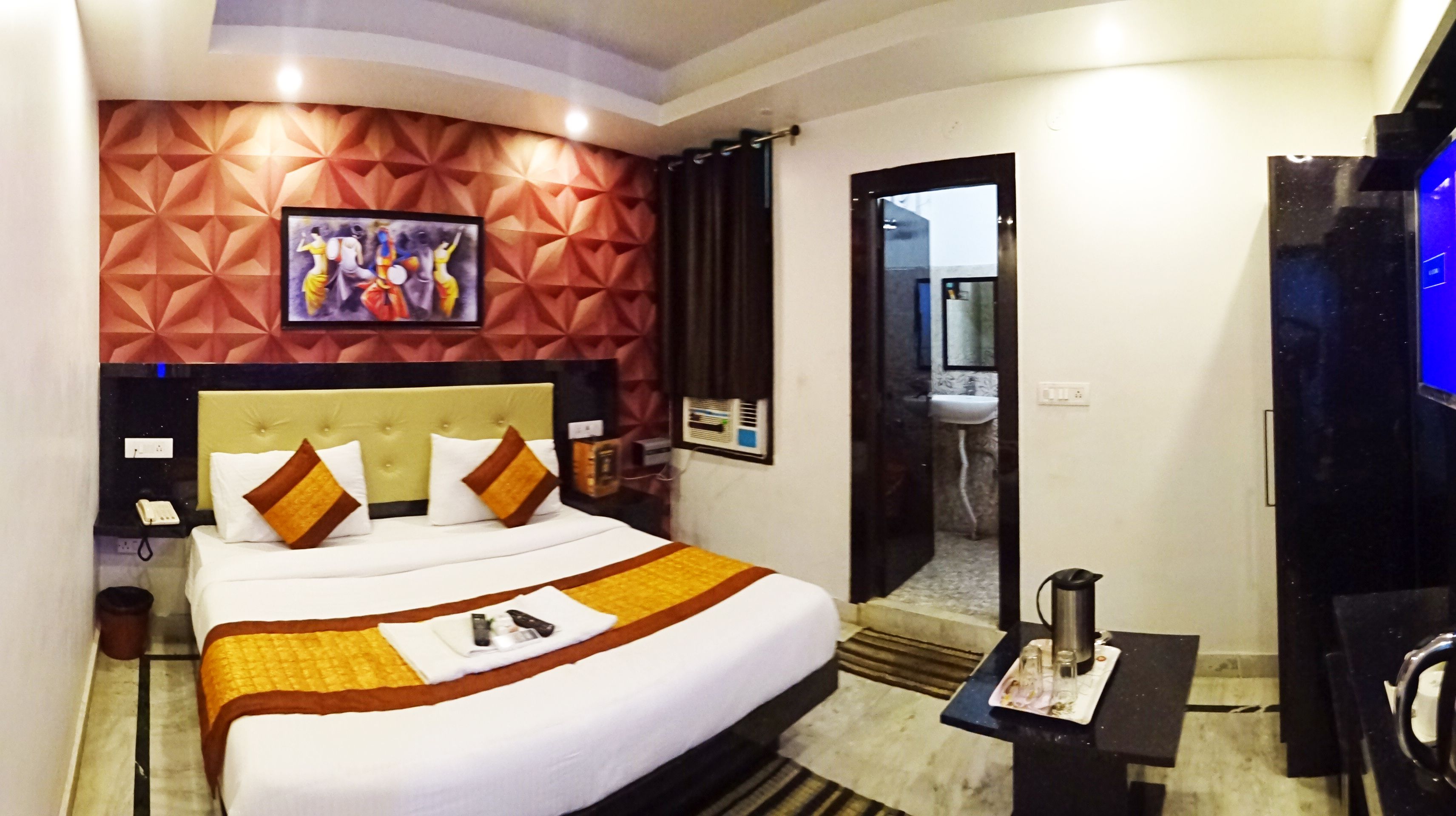 A hotel room with a large bed, a headboard with a geometric pattern, a painting above the bed, a bathroom visible through an open door, and a coffee maker on a small table.