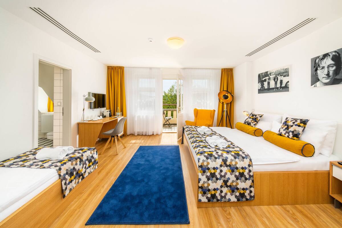 A hotel room with a bed, two bedside tables, a desk, a chair, a blue rug, and a view of the outdoors.