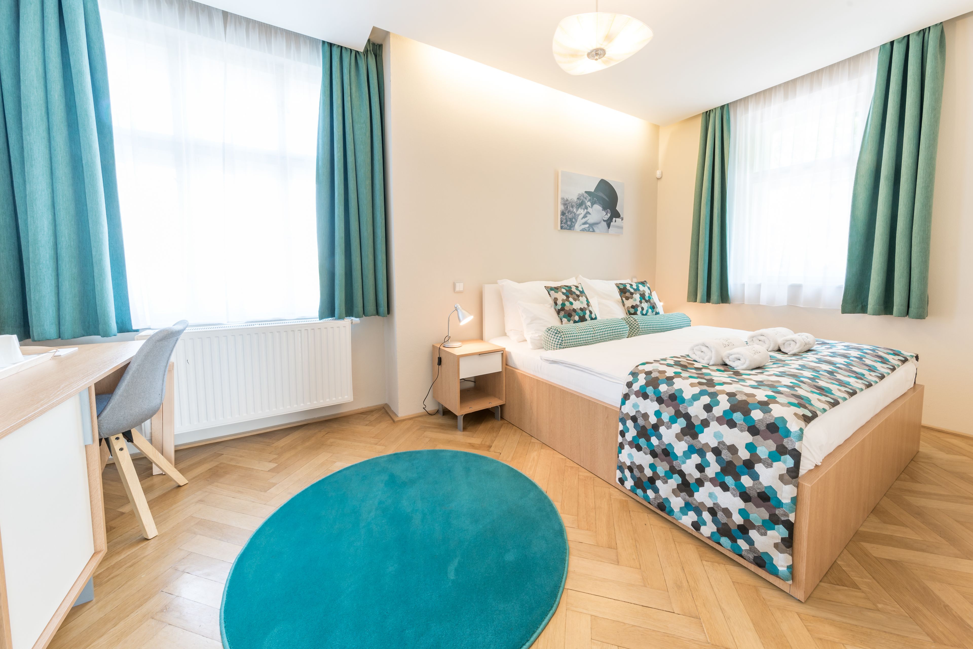 A hotel room with a bed, a desk, a chair, a nightstand, and a large window with curtains.