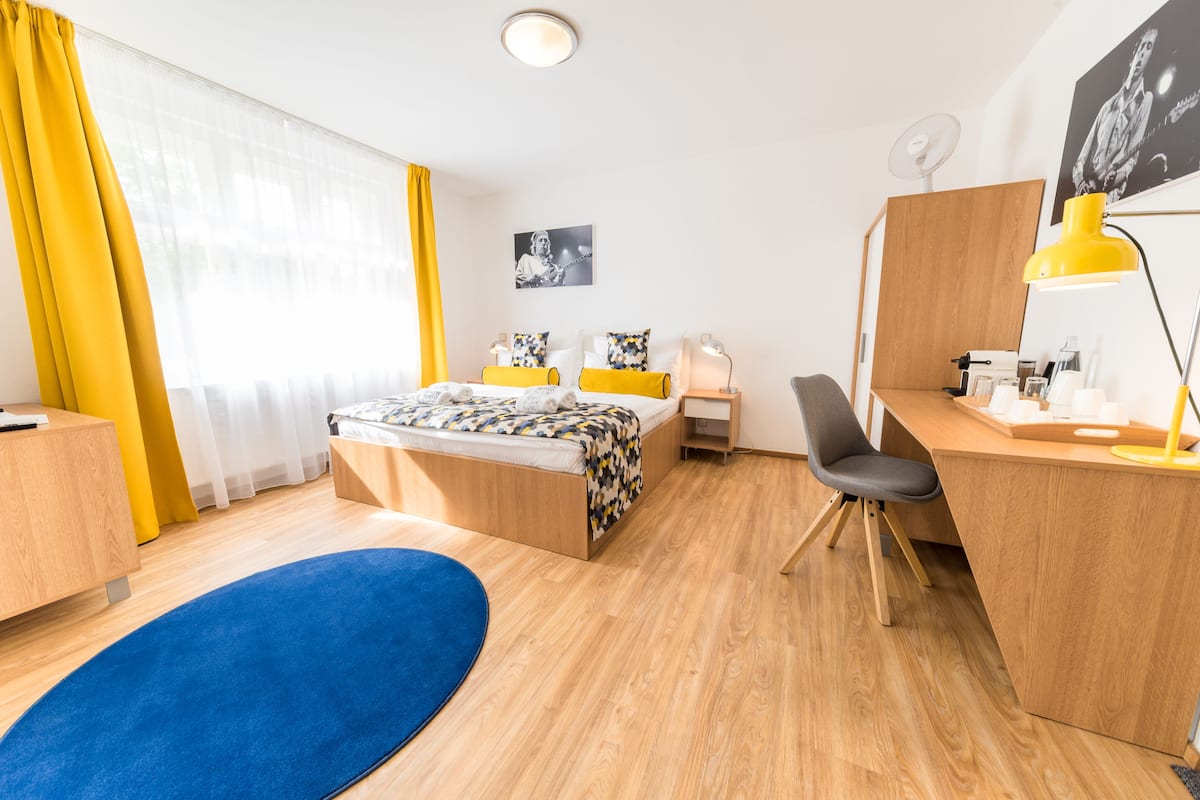 A modern hotel room with a wooden bed, a desk, a chair, a desk lamp, a small table, and a large window with curtains.