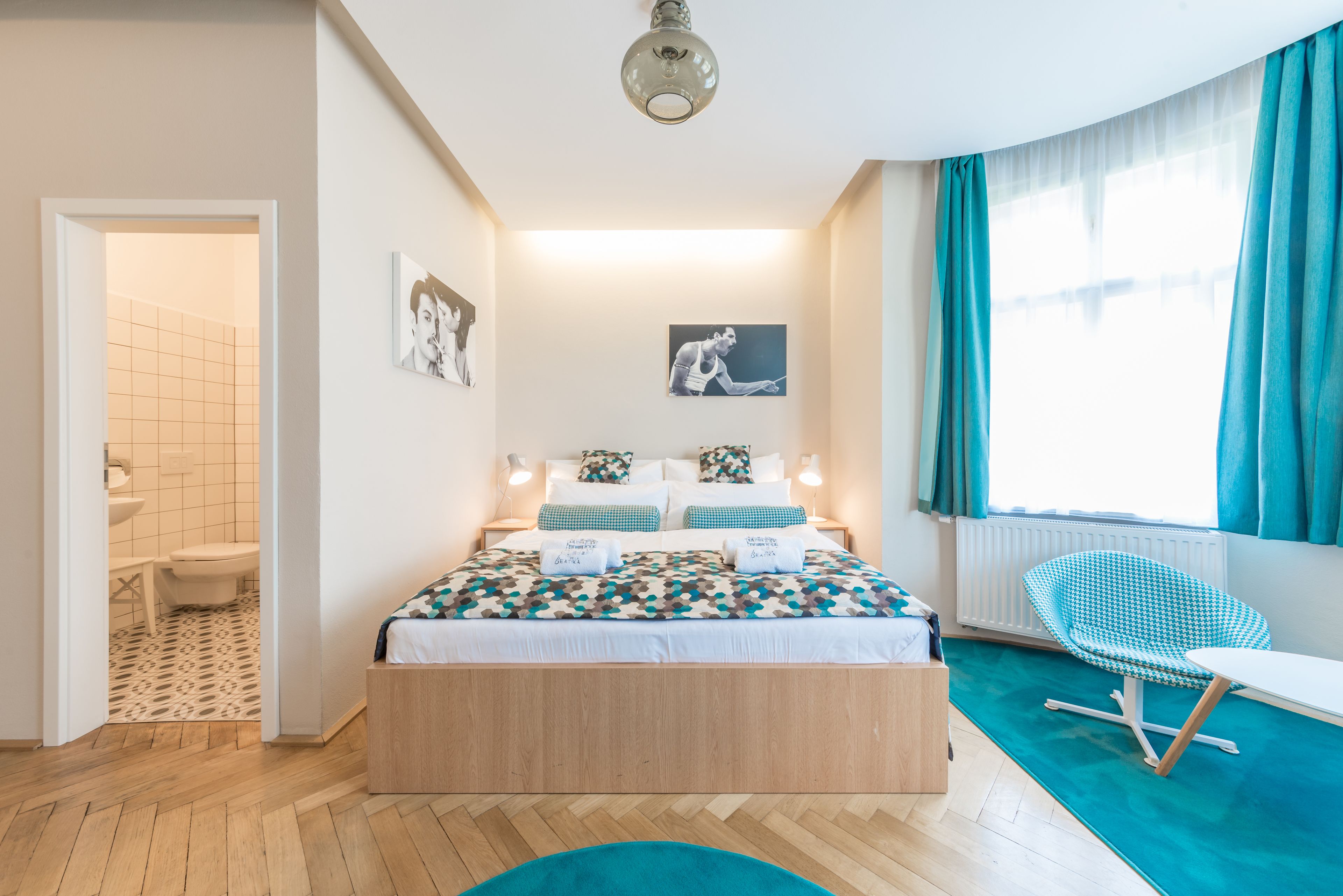Un dormitorio moderno con una cama de madera, una silla azul y vistas al exterior a través de una ventana con cortinas azul turquesa.