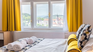 Habitación de hotel con una cama, cortinas amarillas y una ventana con vistas a edificios y colinas.