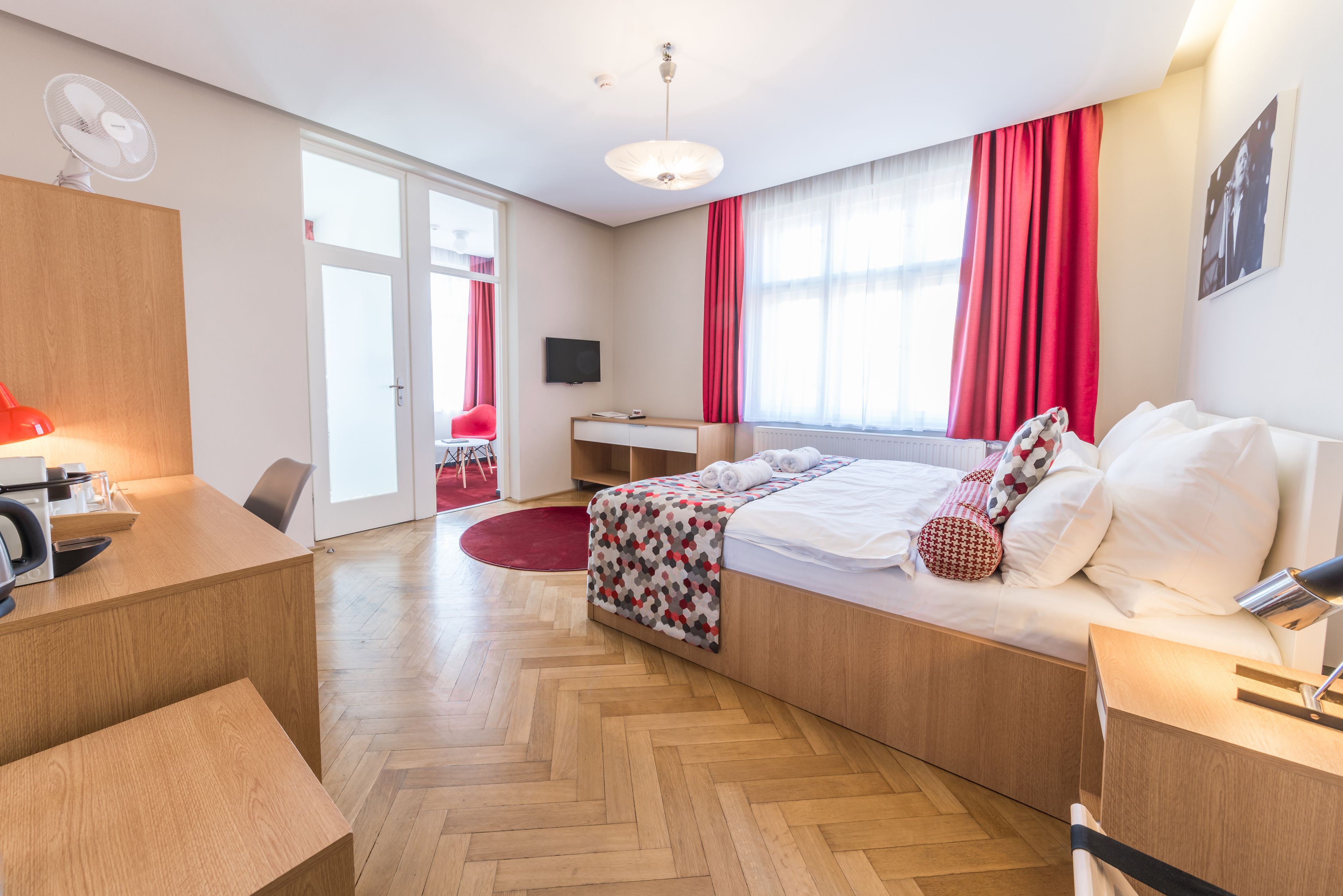 A modern hotel room with a wooden floor, a bed with white bedding, a desk with a chair, a TV, and a red curtain.
