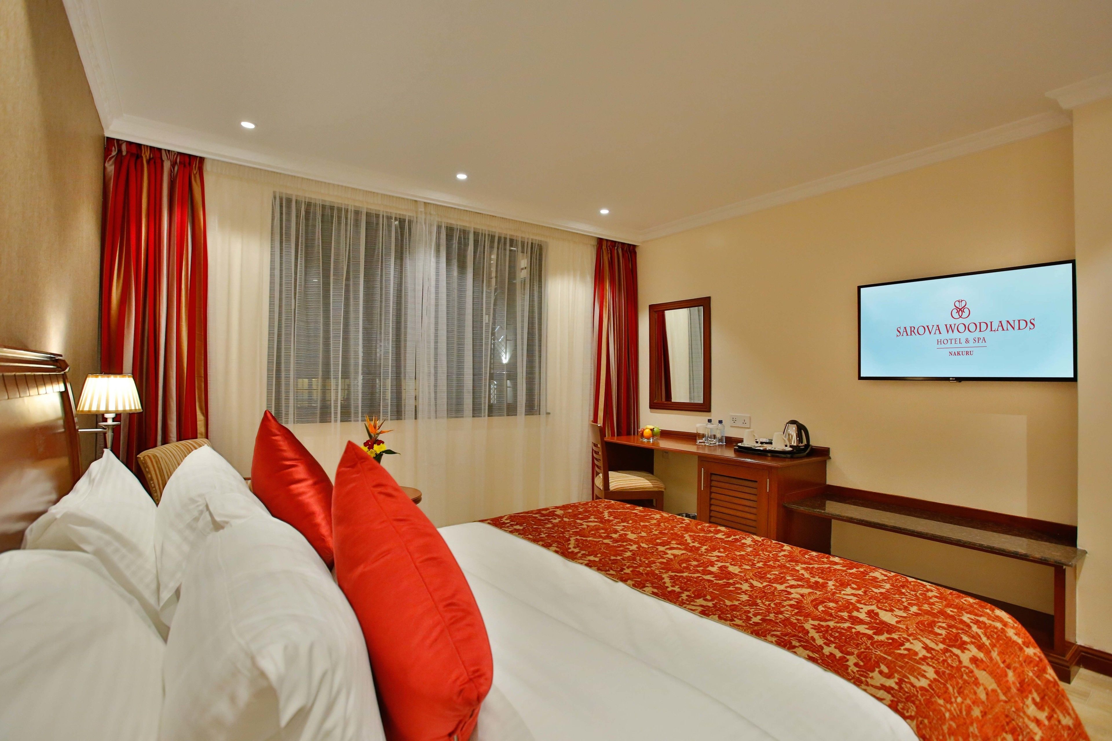 Een hotelkamer met een groot bed, een bureau, een stoel, een televisie waarop de naam van het hotel staat, en een raam met gordijnen.