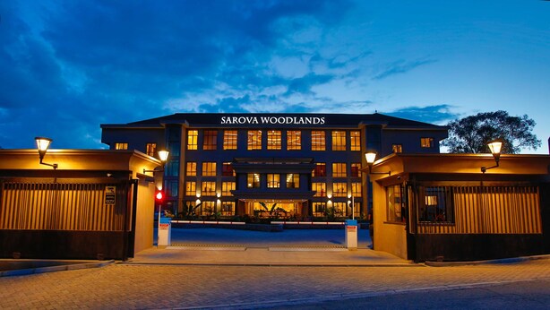 Entrada de hotel con un letrero que dice "SAROVA WOODLANDS", un camino de acceso con portones y una fachada bien iluminada.