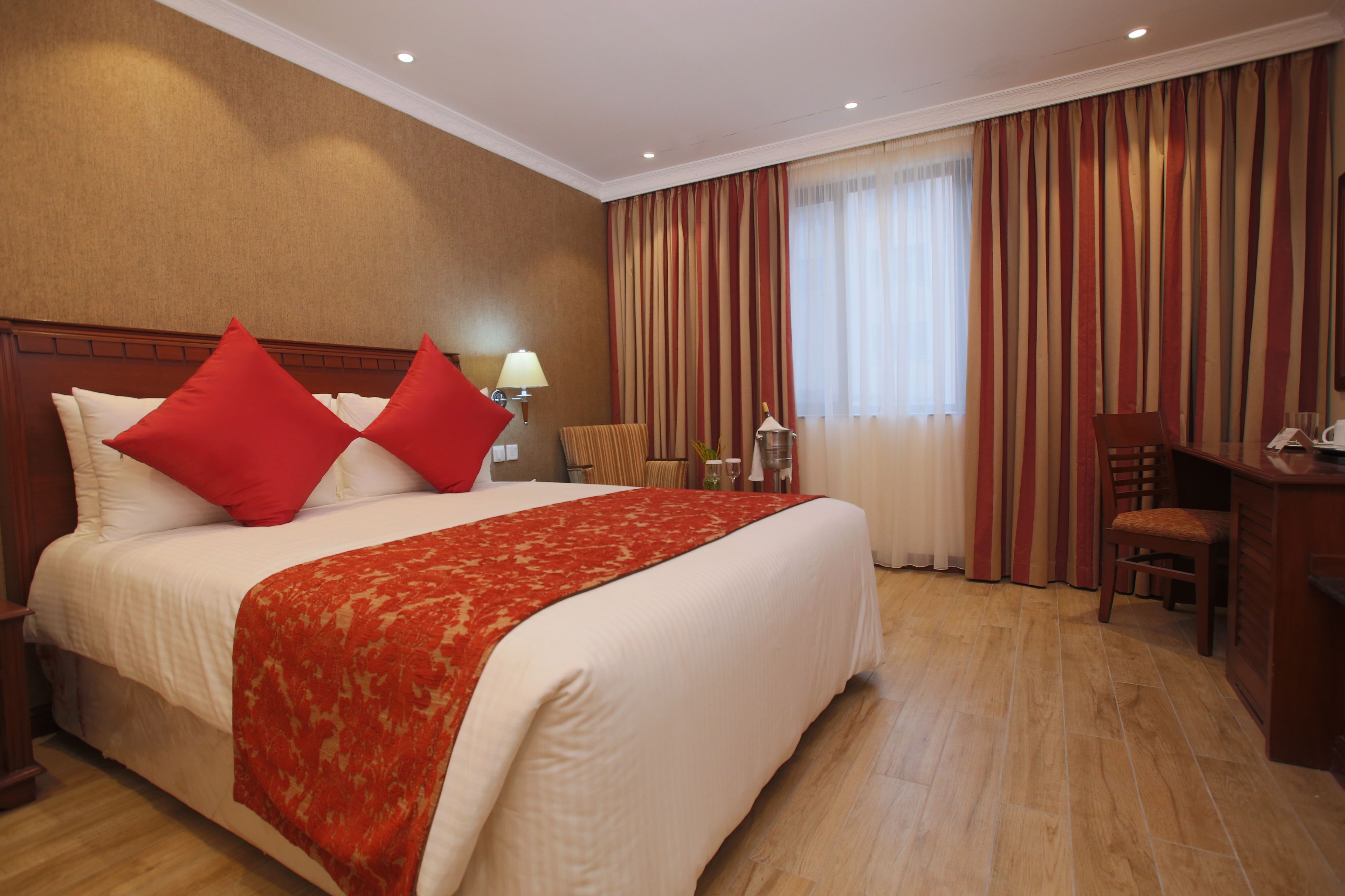 A hotel room with a large bed, red pillows, a red throw blanket, a desk with a chair, and a window with curtains.