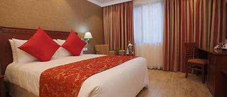 A hotel room with a large bed, red pillows, a red throw blanket, a desk with a chair, and a window with curtains.
