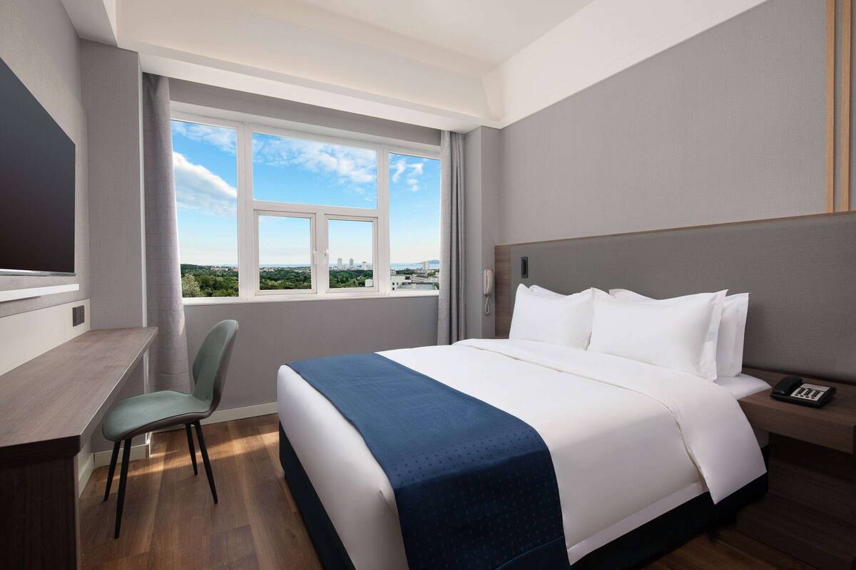 A hotel room with a large bed, a desk, and a chair. There is a window with a view of the city.