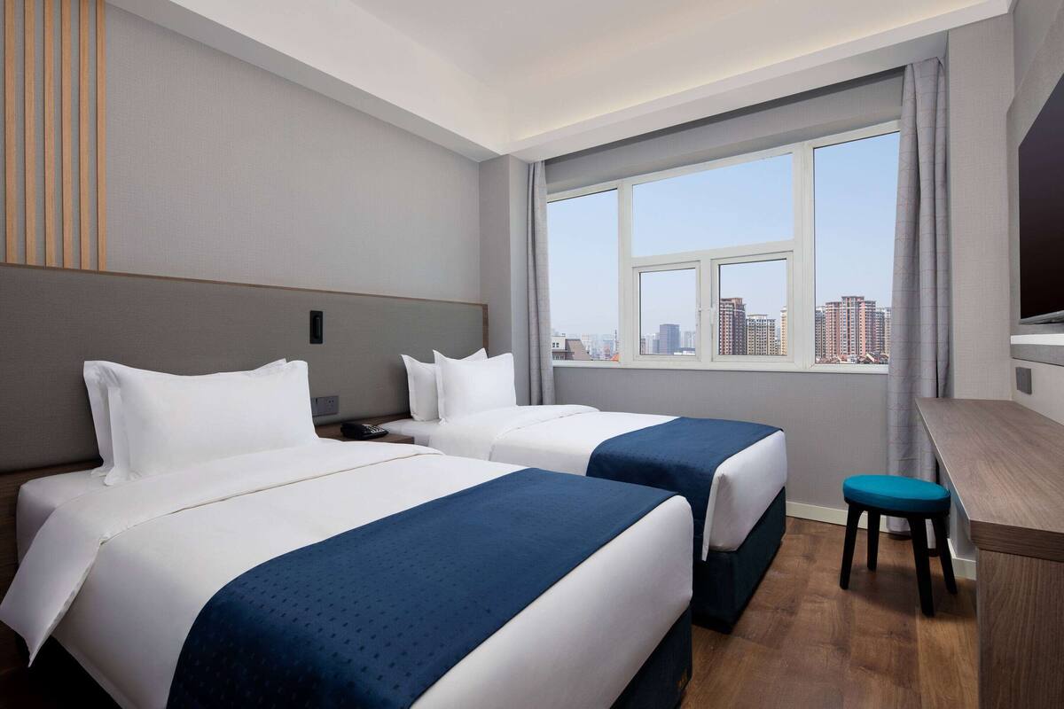 A hotel room with two beds, a desk, a chair, and a large window with a city view.