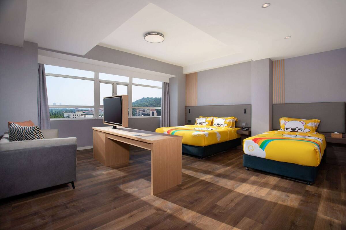 A hotel room with a large window, a desk, two beds with colorful bedding, a sofa, and a television.