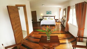 A hotel room with a wooden floor, a bed with white linens, a nightstand, a table with a vase of flowers, and a chair.