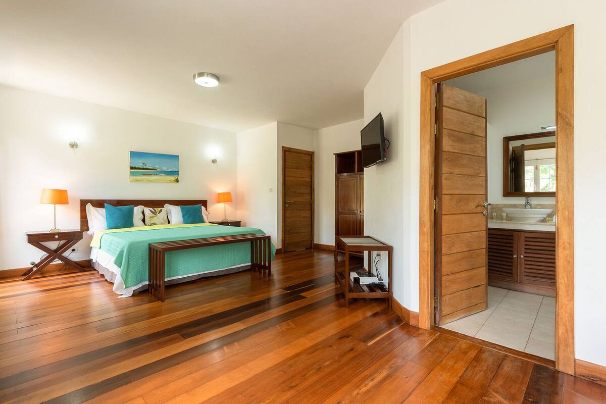 Ein geräumiges Schlafzimmer mit Holzboden, einem Bett mit grüner Bettdecke, zwei Nachttischlampen, einem an der Wand befestigten Fernseher und einer Holztür zu einem Badezimmer mit Waschbecken und Spiegel.