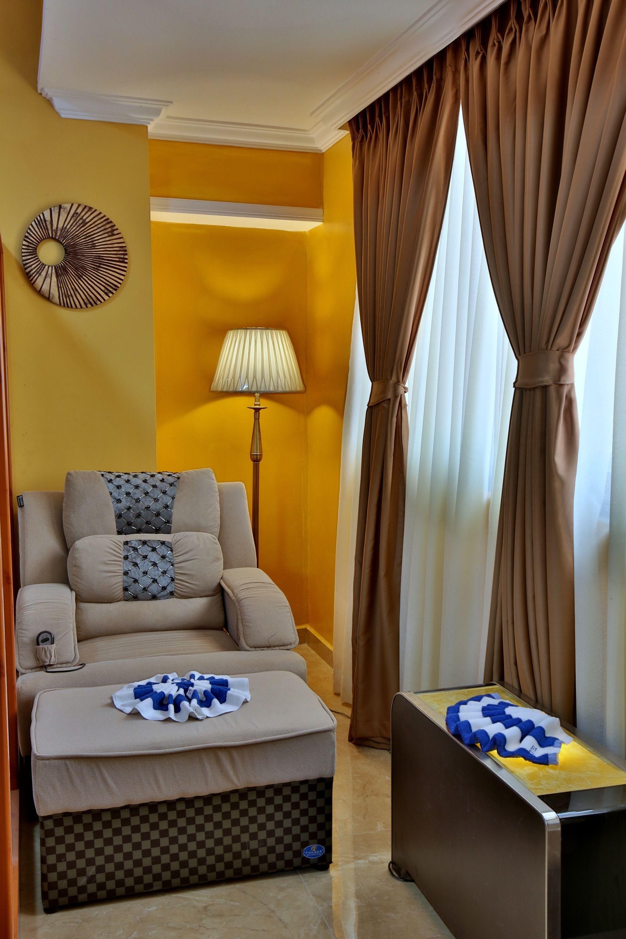 A living room with a sofa, ottoman, and a small table with a blanket. There is a lamp and a wall decoration.