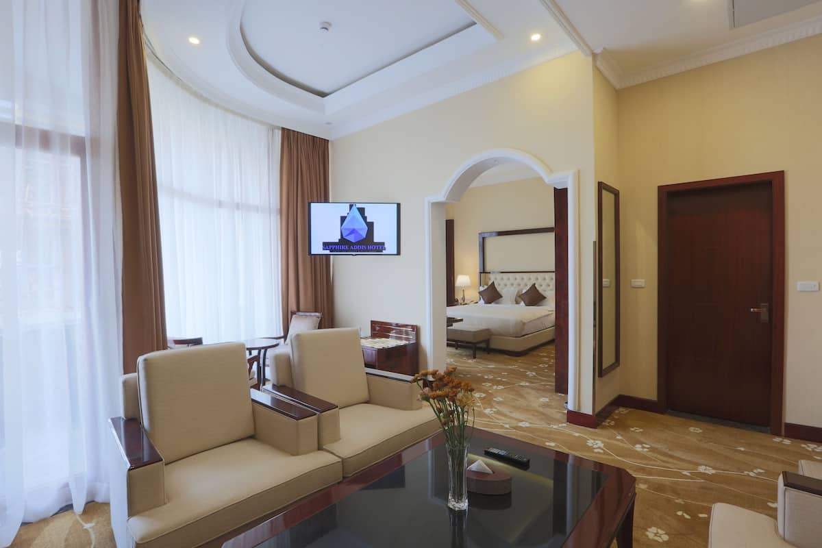A hotel room with a sofa, a coffee table with a vase of flowers, a television, a bed with a headboard, and a door.