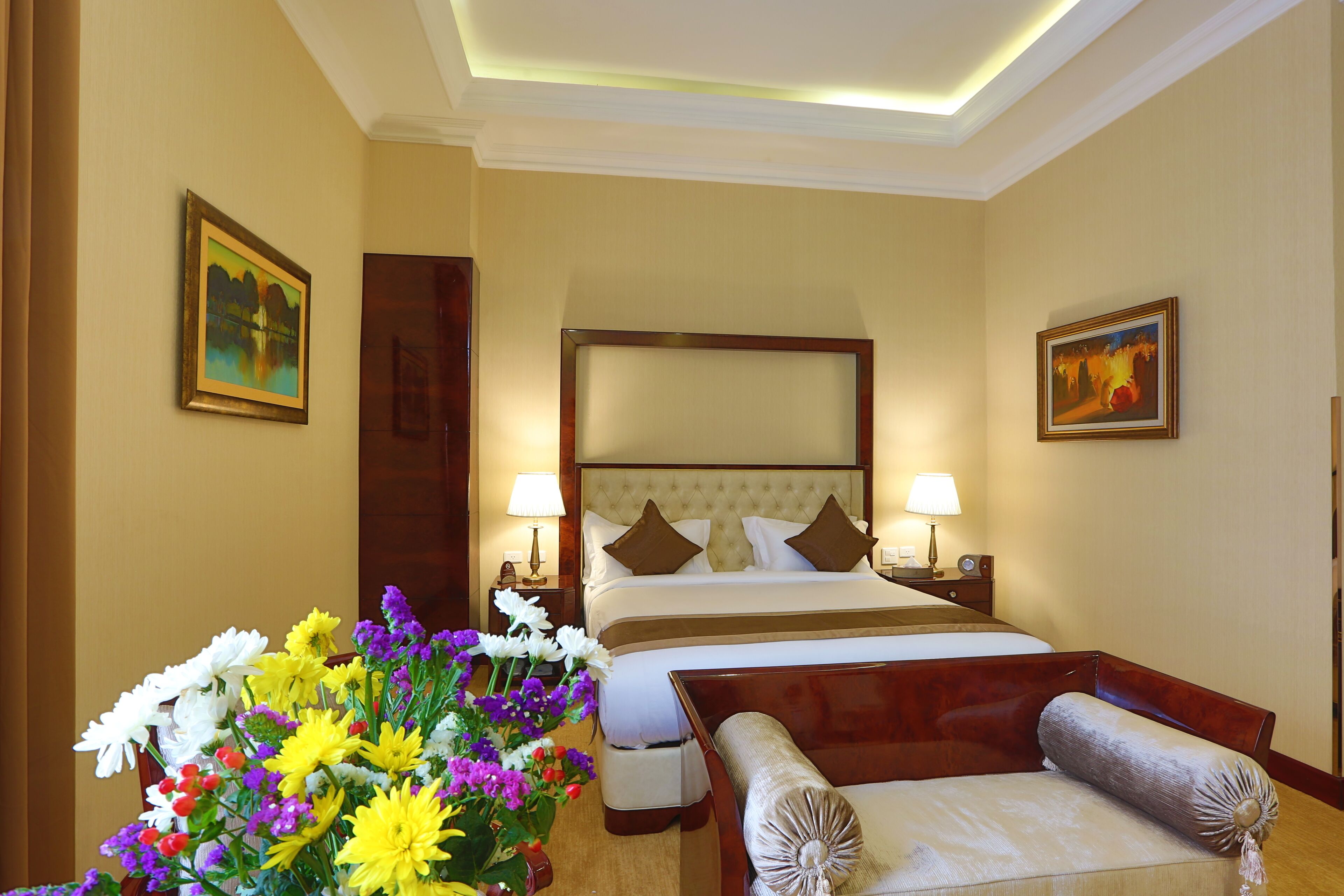 A hotel room with a large bed, two bedside tables with lamps, a wooden chair, and a floral arrangement.