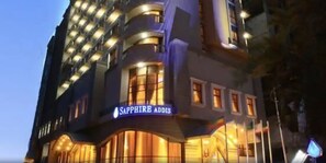 Un hotel de varios pisos con un llamativo letrero 'Sapphire Adonis', grandes ventanales y un diseño arquitectónico moderno.