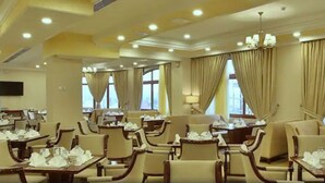 A hotel dining area with multiple tables and chairs, large windows with curtains, and a chandelier.