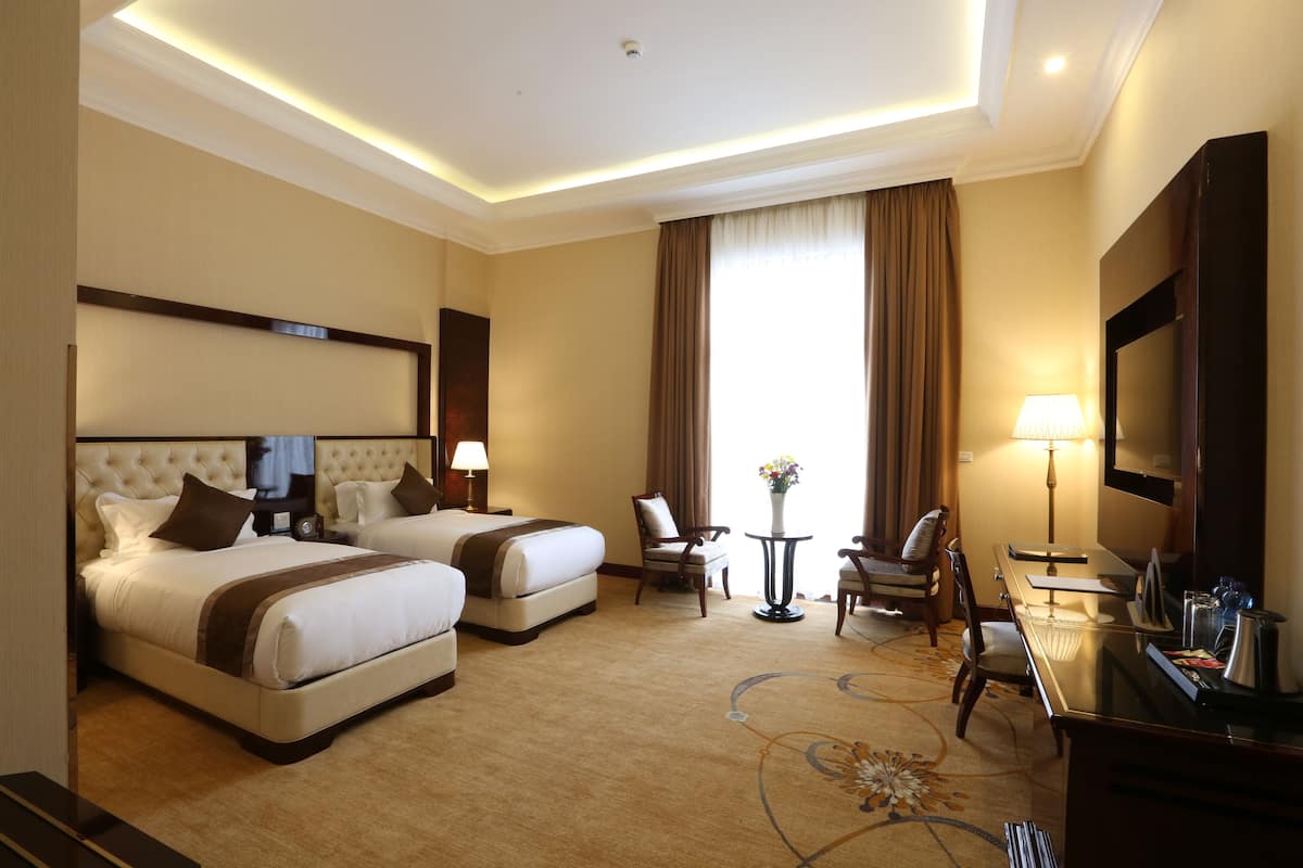 A hotel room with two beds, a desk, a chair, a TV, and a lamp.