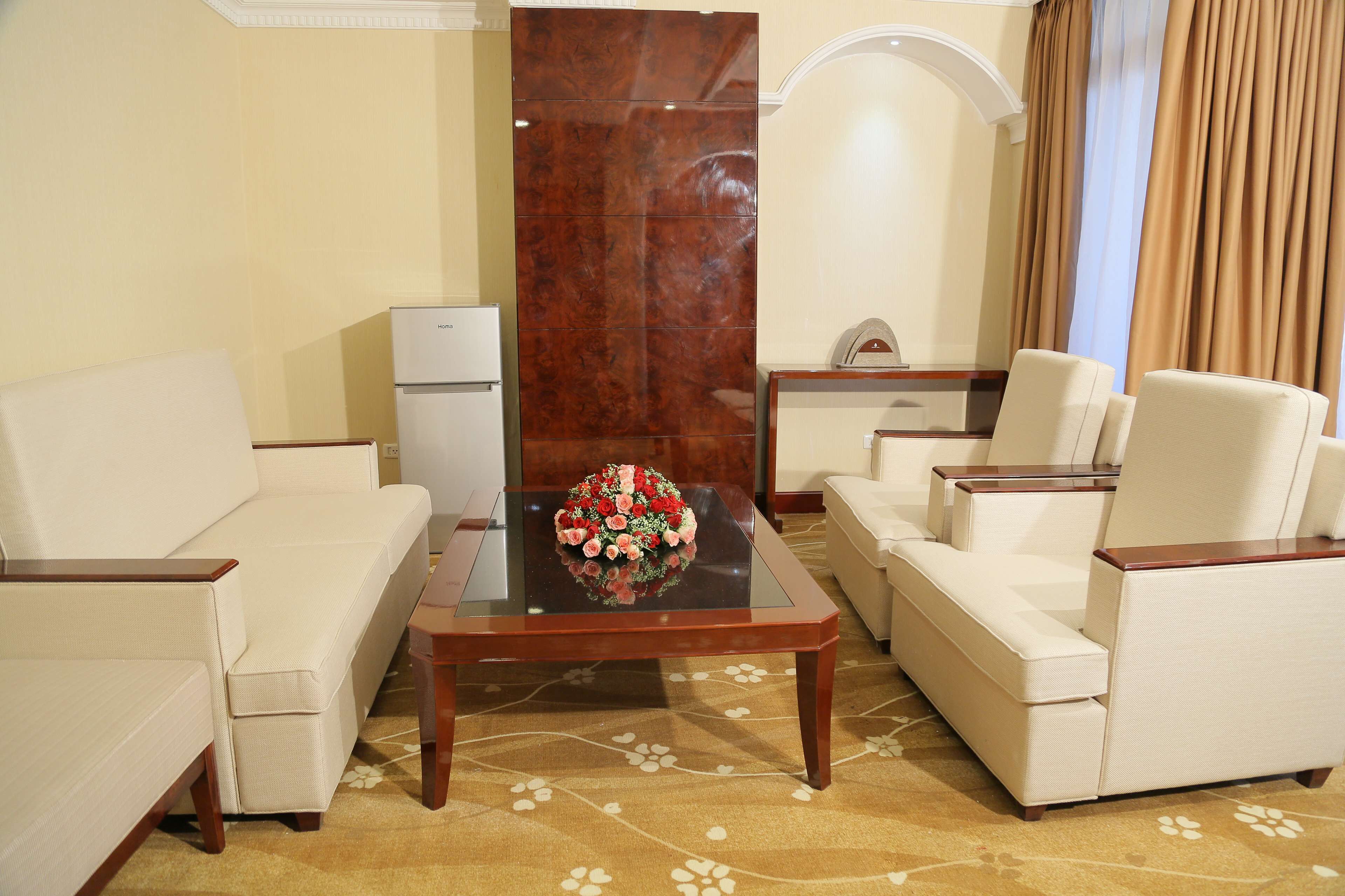 A living room with a sofa, armchairs, a coffee table with a floral centerpiece, a small table with a clock, and a refrigerator.