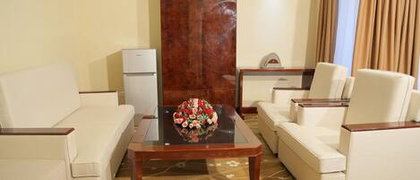 A living room with a sofa, armchairs, a coffee table with a floral centerpiece, a small table with a clock, and a refrigerator.