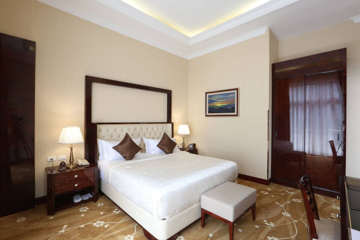 A hotel room with a large bed, two bedside tables with lamps, a wooden wardrobe, and a painting on the wall.