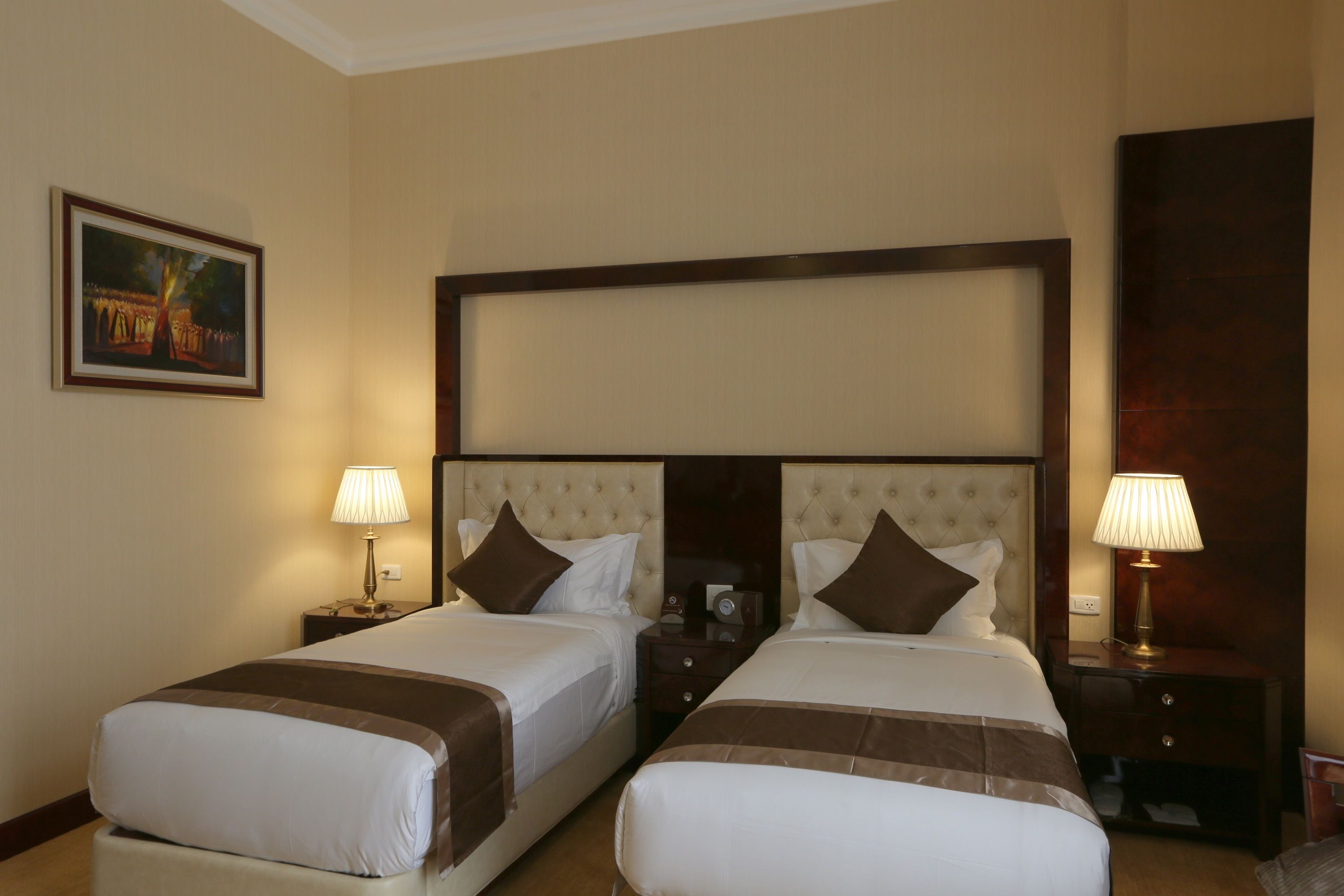A hotel room with two beds, each with a headboard, bedside tables, lamps, and a painting on the wall.