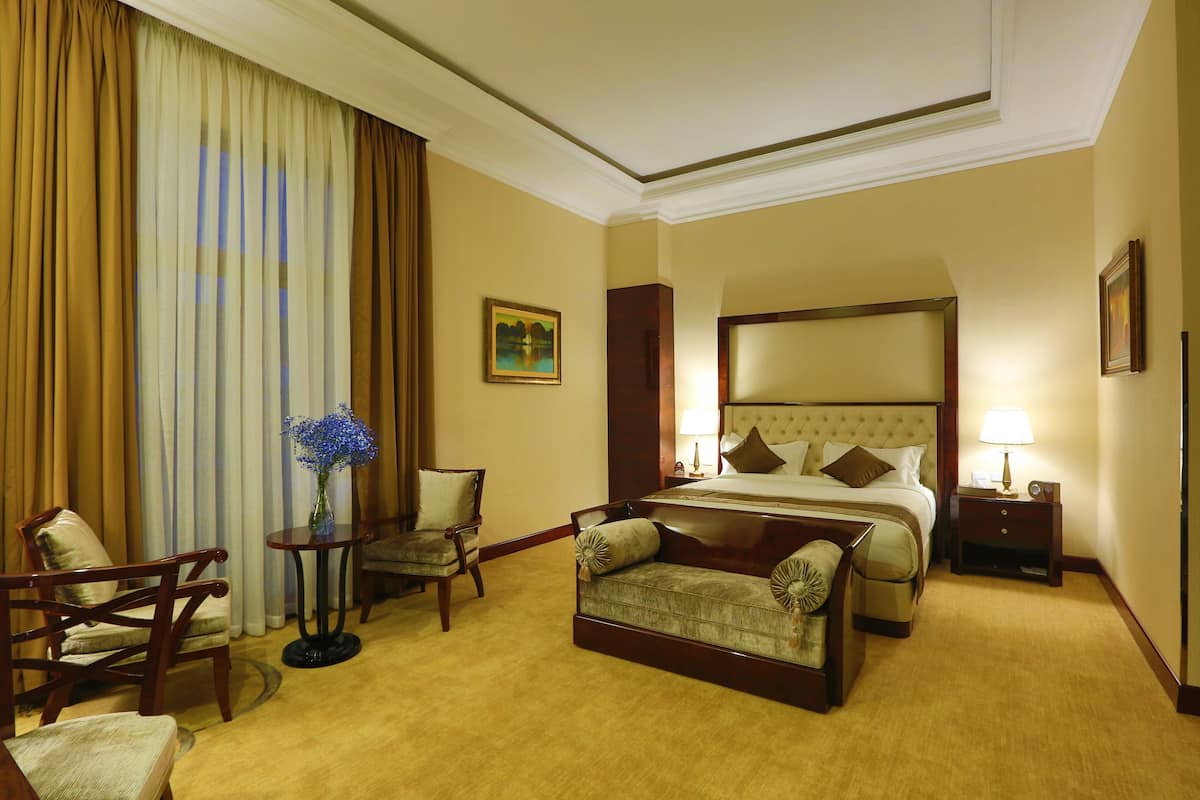 A hotel room with a large bed, a sitting area with a sofa and chairs, a small table with a vase, and a painting on the wall.