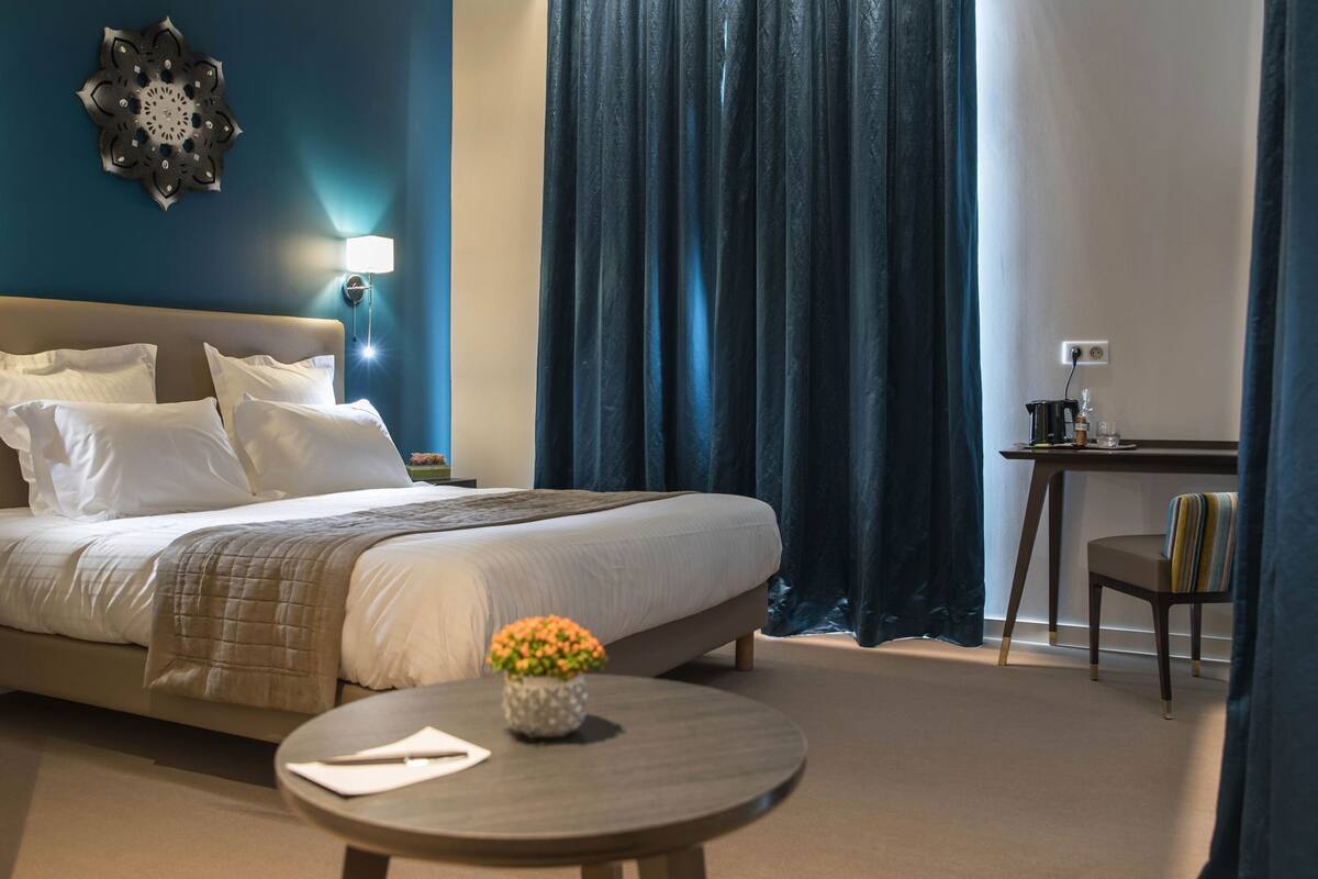 A neatly made bed with pillows, a bedside table with a lamp, a small round table with a vase of flowers, and a desk with a cup and glass.