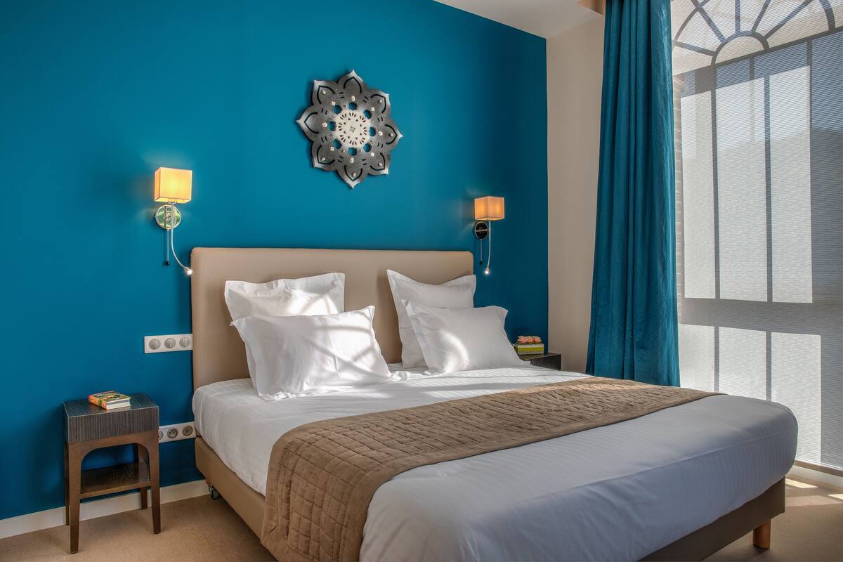 A neatly made bed with a beige headboard and white bedding, two wall-mounted lamps, a decorative wall piece, and a window with blue curtains.