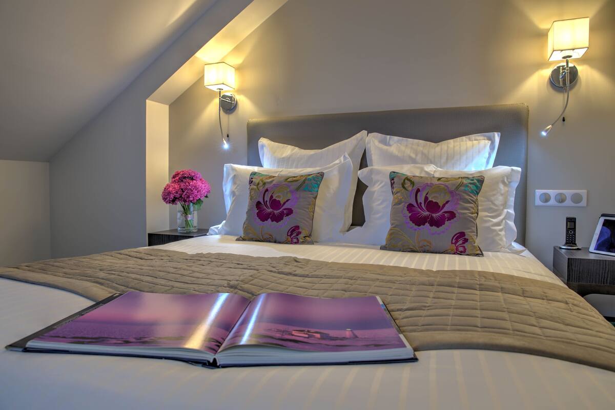 A neatly made bed with a quilted cover, two wall-mounted lamps, and a bedside table with a phone and a small vase with flowers.