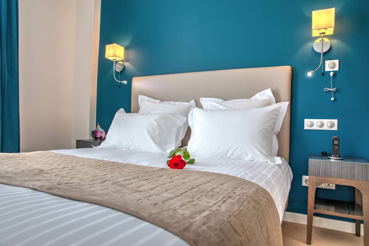 A neatly made bed with white linens, a beige throw blanket, and a single red rose.