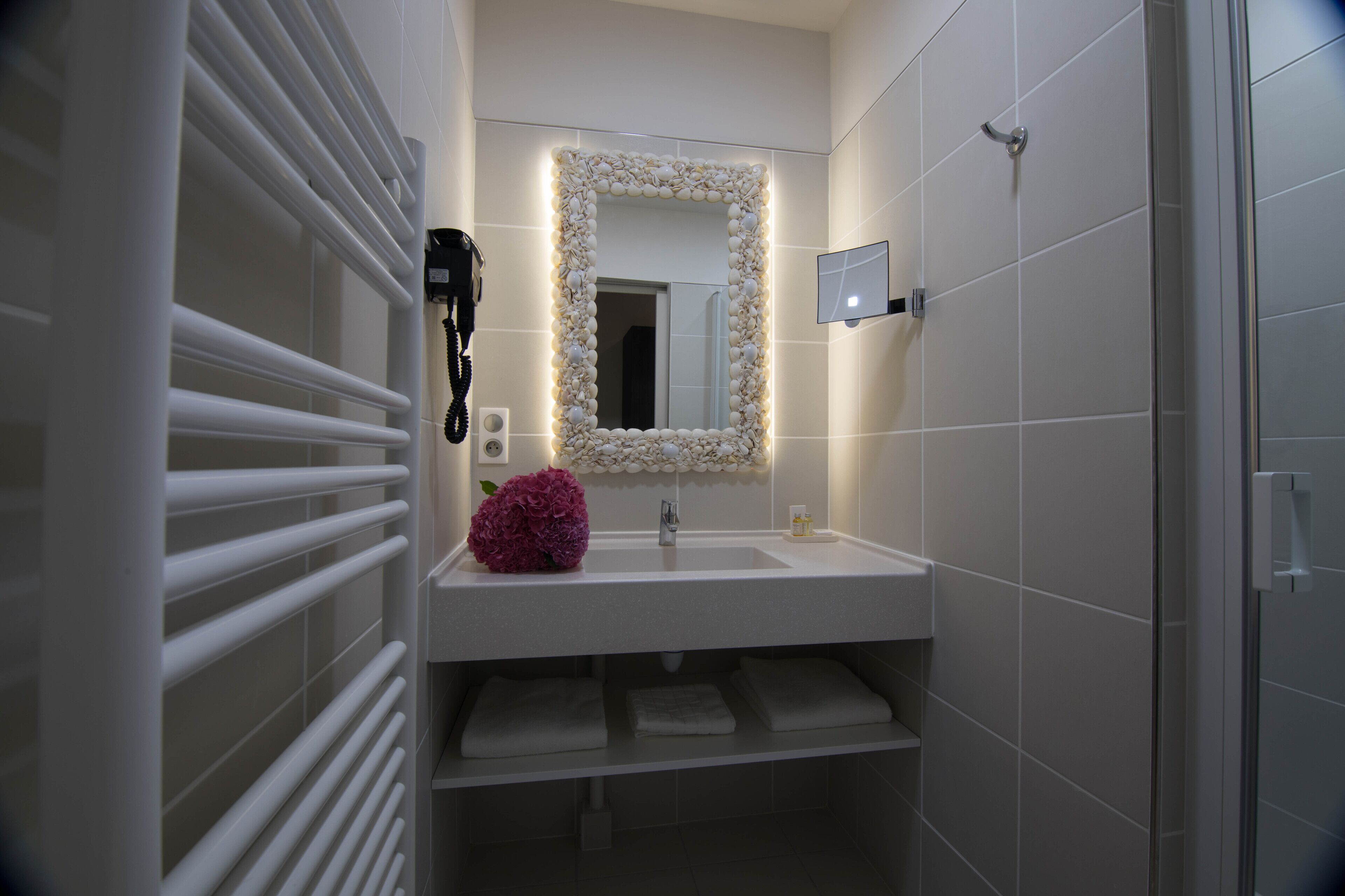 A modern bathroom with a white sink, a mirror with a decorative frame, a hairdryer, and a pink flower.