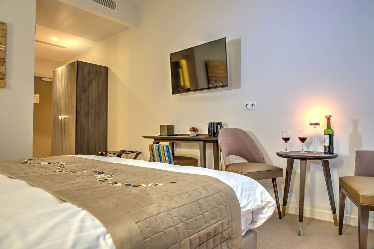 A hotel room with a bed, a desk, two chairs, a table with wine glasses and a bottle, and a large mirror on the wall.