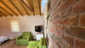Una habitación con techo de madera, pared de ladrillo, sofá verde, una mesita, cama y televisión.