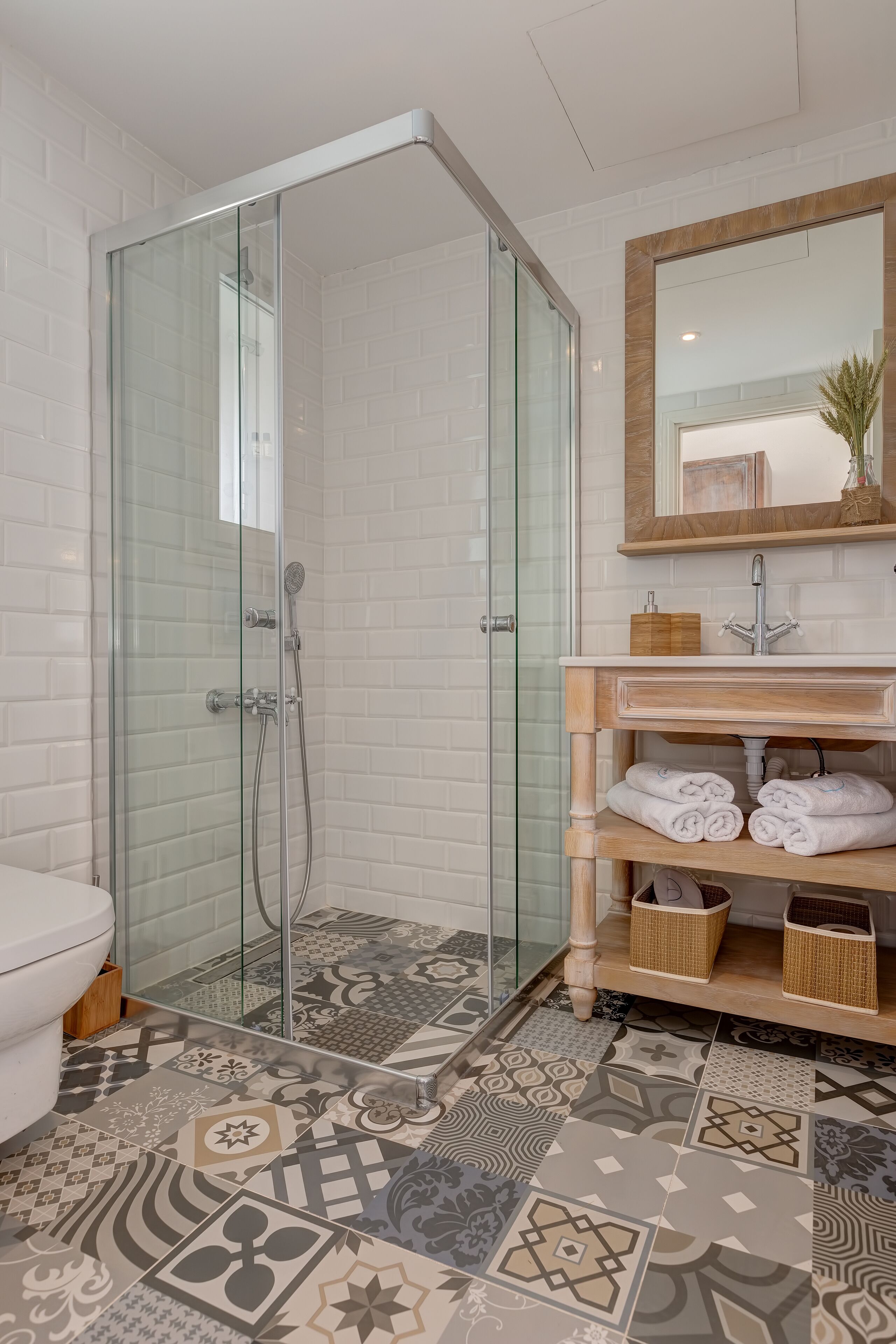 A modern bathroom with a glass-enclosed shower, a white toilet, a wooden vanity with a mirror, and patterned floor tiles.