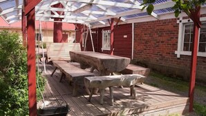 Ulkotilaan sijoitettu puinen terassi, jossa on pergola, kivipöytä ja penkkejä.