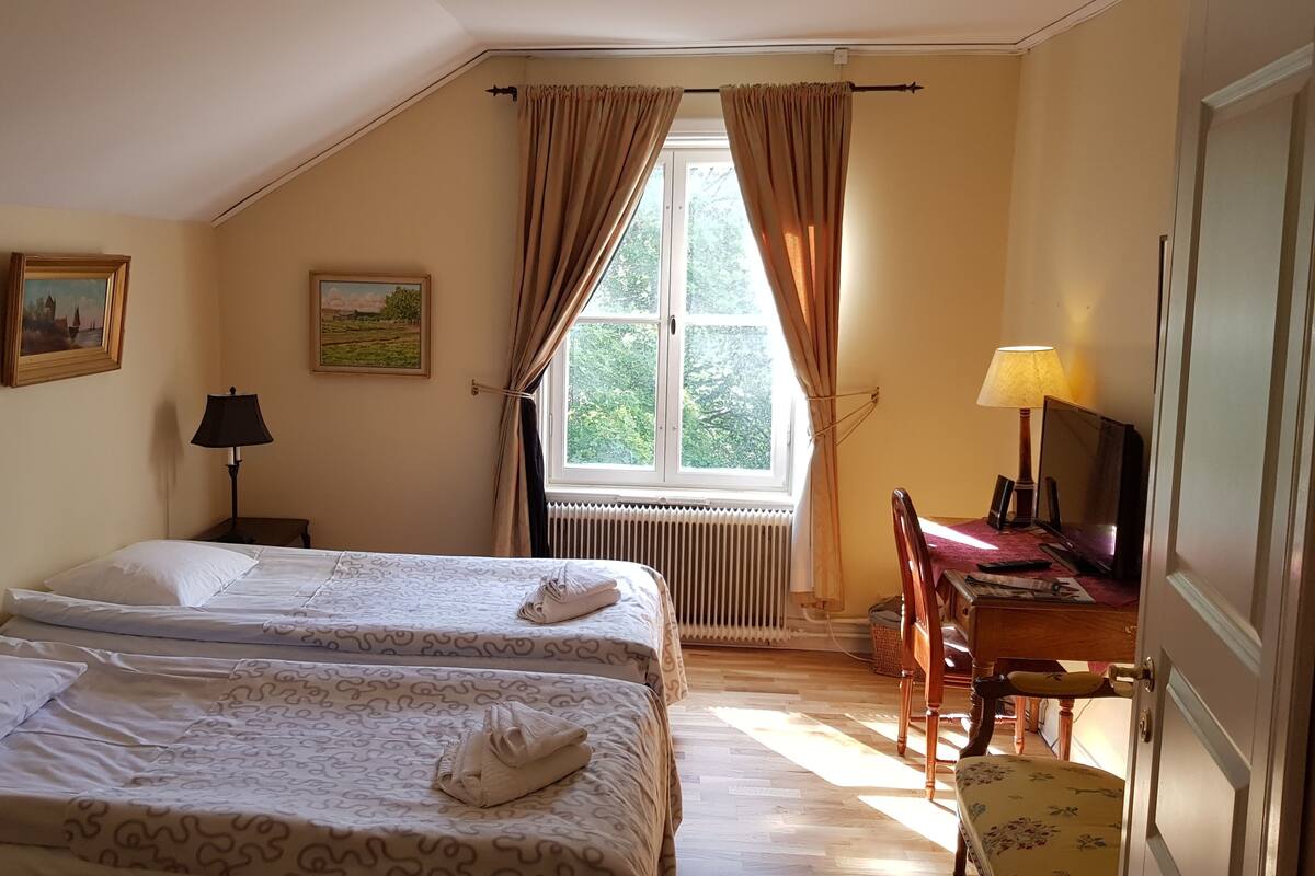 Habitación de hotel con dos camas, un escritorio con lámpara, una silla y un ventanal con cortinas.