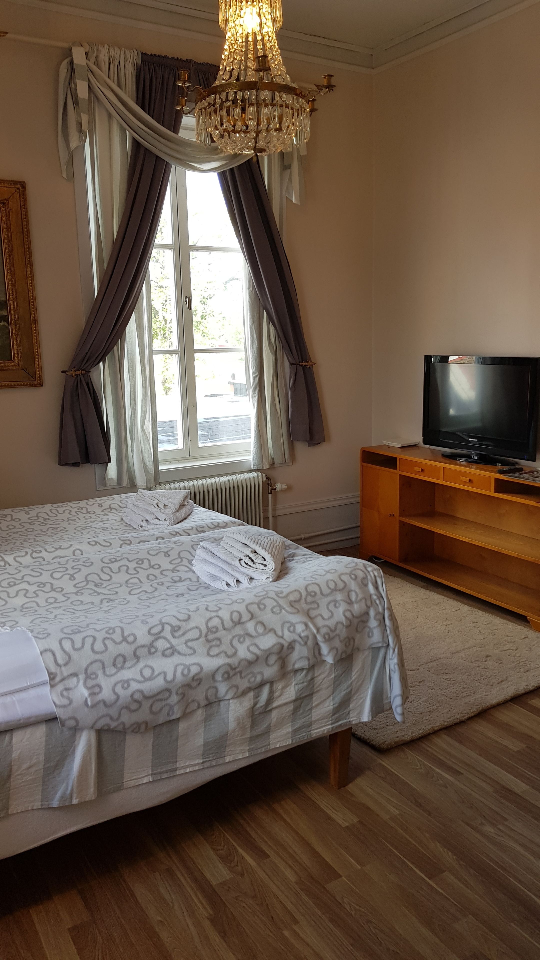 A neatly made bed with a patterned bedspread, a television on a wooden stand, and a chandelier.